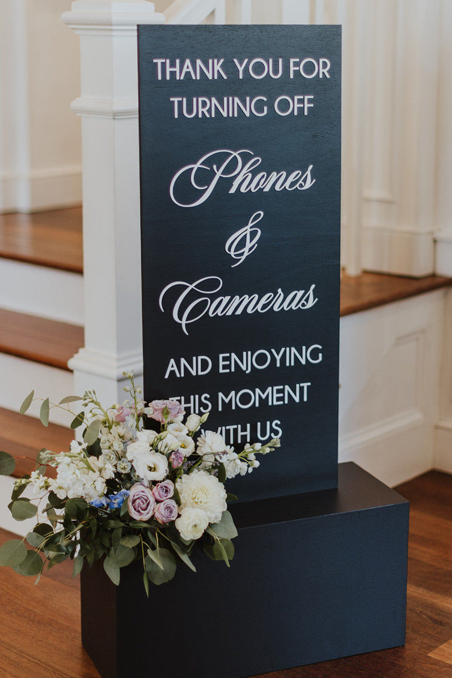 Decorative sign asking guests to turn off phones and cameras with a floral arrangement.